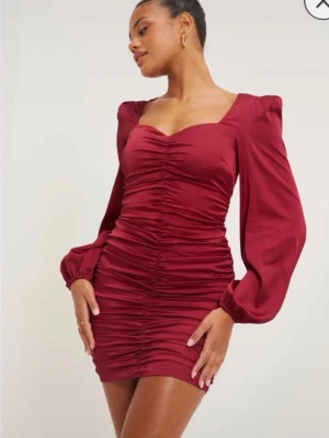 Vinröd omlottklänning med puffärm - Supersnygg vinröd kort klänning med långa puffärmar och omlottdetalj. Klänningen har rynkade sömmar framtill som ger en smickrande siluett och en feminin v-ringning. Tillverkad i ett glansigt polyestertyg som känns lyxigt.