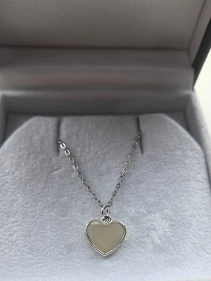 Halsband med självlysande hjärthänge - Snyggt halsband med ett hjärtformat hänge som lyser i blått i mörker och är vitt/creme/svagt glittrig i dagsljus. Kedjan är i rostfritt stål och hänget har en slät, glansig yta. Perfekt för dig som gillar unika accessoarer med cool effekt. Hjärtat började lysa i mörkare omgivning efter att jag tog kort på det i fönstret. (Vanligt dagsljus, även molnigt väder, aktiverar skenet i hänget).