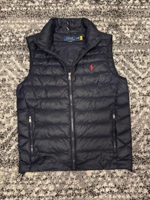 Mörkblå dunväst från Polo Ralph Lauren - Snygg mörkblå dunväst från Polo Ralph Lauren med klassisk quiltad design och röd broderad logga på bröstet. Västen har två fickor med dragkedja och hög krage. Perfekt för lager på lager och riktigt stilren till hösten. Västen är i storlek xs men passar framför i S. Ny pris kostar den 3000. Hör av er vid eventuella frågor eller funderingar!!