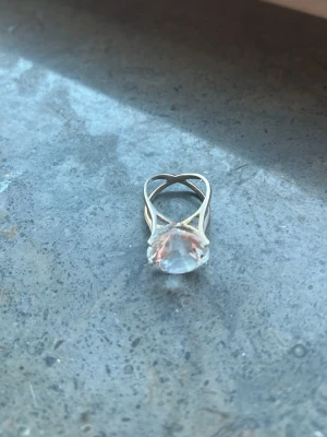 Guldig ring med stor klar sten - Unik ring i guldfärgad metall med en stor, klar slipad sten som centerpiece. Ringen har en modern, korsad design som ger ett coolt och trendigt intryck. Perfekt för dig som vill sticka ut med ett statement-smycke.