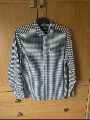 Rutig skjorta från Lyle & Scott - Snygg rutig skjorta från Lyle & Scott i blått och vitt med klassisk krage och gul logga på bröstet. Skjortan har långa ärmar och knäppning framtill. Perfekt för en clean och stilren look.