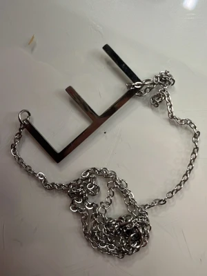 Silverfärgat halsband med geometriskt hänge - Säljer ett halsband i silverfärgad metall med ett stort, geometriskt hänge i form av tre vinklade linjer. Kedjan är tunn och har en enkel länkdesign. Perfekt för dig som gillar minimalistiska och moderna accessoarer.