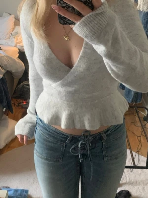 Ljusgrå omlottstickad tröja - Säljer en ljusgrå stickad tröja med omlottmodell och djup v-ringning. Tröjan har långa ärmar och en volangkant nedtill som ger en feminin touch. Mjuk och skön, perfekt till jeans eller kjol.