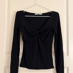 Svart långärmad topp från NA-KD - Trendig svart långärmad topp från NA-KD med djup v-ringning och twistad detalj framtill. Figurnära passform och mjukt stretchigt material som sitter snyggt på kroppen. Användt den några gånger 