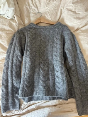 Grå kabelstickad tröja - Mysig grå stickad tröja med kabelstickat mönster och rund hals. Tröjan har långa ärmar och ribbade muddar vid ärmslut och nederkant. Perfekt för kyliga dagar och enkel att matcha med jeans eller kjol.