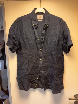 Mörkblå linneskjorta från Knowledge Cotton - Snygg mörkblå skjorta i linne från bläck. Skjortan har korta ärmar, klassisk krage och knappar framtill. Perfekt för varma dagar och ger en avslappnad men stilren look.