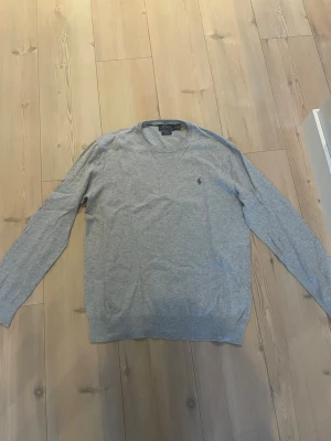 Grå stickad tröja Polo Ralph Lauren - Snygg grå stickad tröja från Polo Ralph Lauren i slim fit-modell. Tillverkad i mjuk pima cotton och har rund halsringning samt diskret logga broderad på bröstet. Perfekt för en stilren och enkel look.