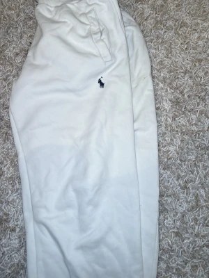Vita mjukisbyxor från Polo Ralph Lauren - Säljer ett par vita mjukisbyxor från Polo Ralph Lauren med dragsko i midjan och två sidofickor. Byxorna har den klassiska blå loggan broderad på vänster sida och är gjorda i mjukt bomullsmaterial. Perfekta för en chill och avslappnad stil.