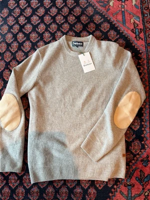Grå stickad tröja från Barbour - Snygg grå stickad tröja från Barbour med rund halsringning och ribbade muddar. Tröjan har beige mockadetaljer på armbågarna som ger en klassisk touch. Perfekt för dig som gillar stilrena och tidlösa plagg.