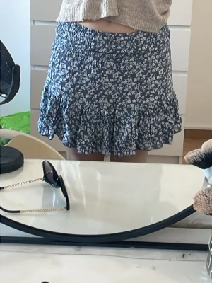 Blommig volangkjol i blått och vitt - Supersöt kort kjol med blå botten och vitt blommönster. Kjolen har bred resår i midjan och volangkant nedtill som ger en flowy känsla. Perfekt för sommardagar och enkel att matcha med en crop top eller stickad tröja. Materialet känns lätt och luftigt.