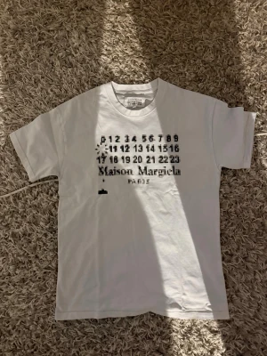 Maison Margiela vit t-shirt med tryck - Vit t-shirt från Maison Margiela med svart grafiskt tryck och logga på bröstet. Klassisk passform med rund hals och korta ärmar. Tillverkad i mjuk bomull som känns skön mot huden. Perfekt för dig som gillar minimalistisk och modern stil.