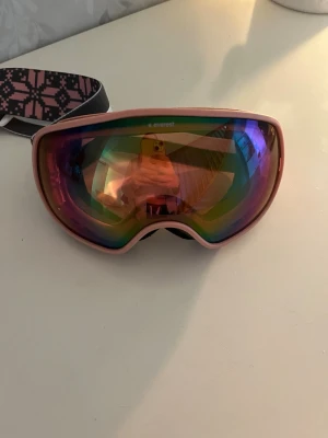 Everest skidglasögon rosa spegel - Säljer ett par snygga skidglasögon från Everest med rosa båge och spegelglas i regnbågsfärger. Bred elastisk rem med gråt och rosa mönster samt Everest-logga. Perfekta för skidåkning eller snowboard. Medföljer vit förvaringspåse. Liten fläck på den vita påsen. 