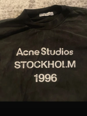 Svart Acne Studios t-shirt - Säljer en svart t-shirt från Acne Studios med vit text 'Acne Studios STOCKHOLM 1996' på bröstet. Klassisk rund hals och korta ärmar. Perfekt för dig som gillar minimalistisk och stilren design.