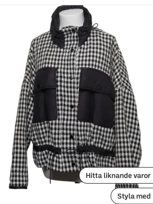 Rutig svartvit vindjacka med snörning - Snygg vindjacka i svartvitt rutigt mönster med stora svarta fickor framtill och snörning i midjan. Jackan har hög krage, knappstängning och långa ärmar. Xs-M