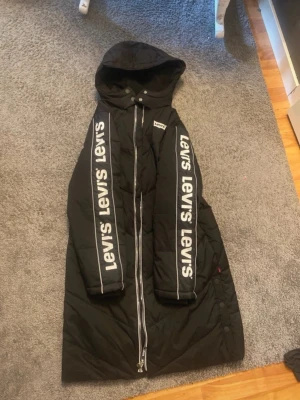 Svart lång dunjacka från Levi's - Säljer en svart lång dunjacka från Levi's med huva och dragkedja framtill. Jackan har stora vita Levi's-loggor längs båda ärmarna och en logga på bröstet. Snygg streetstil och perfekt för kalla vinterdagar.