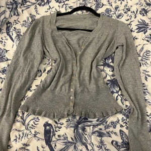 Grå kofta med volangkanter! - Säljer en ljusgrå kofta med långa ärmar och v-ringning. Koftan har ribbade muddar, volangkant nedtill och knäpps med små vita knappar framtill. Säljer pågrund av att den inte passar. (Skriv för frågor eller prisförslag med mera!)💕