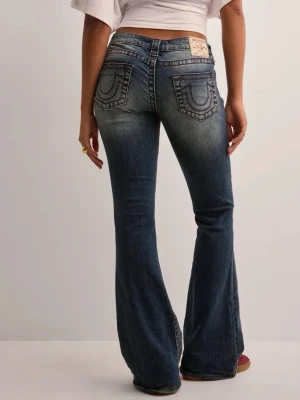 Blå bootcut jeans från True Religion - Säljer ett par blå bootcut jeans från True Religion med klassiska kontrastsömmar och broderade bakfickor. Jeansen har låg midja och är tillverkade i denim med lätt tvättad effekt. Perfekta för en avslappnad och trendig look.