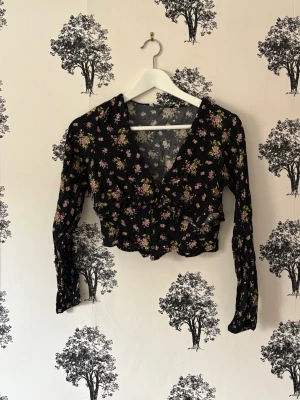 Blommig croppad blus med volang - Snygg svart croppad blus med blommigt mönster i lila, gult och grönt. Djup v-ringning, långa ärmar och volangdetalj framtill. Perfekt för dig som vill ha en trendig och feminin topp med romantiska vibbar.