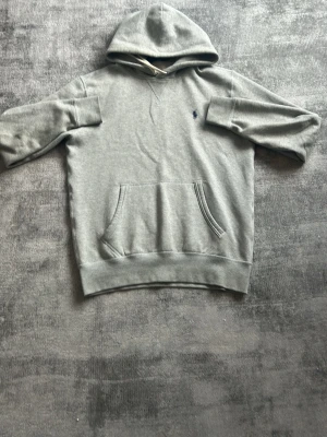 Grå hoodie från Polo Ralph Lauren - Snygg grå hoodie från Polo Ralph Lauren med klassisk huva och känguruficka framtill. Liten mörkblå logga broderad på bröstet. Tillverkad i mjukt bomullsmaterial som är skönt att ha på sig. Perfekt för en avslappnad och stilren look.