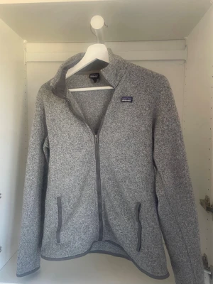 Grå fleecejacka från Patagonia - Säljer en riktigt snygg och tidlös fleece från Patagonia i en gråmelerad färg som passar till allt. Perfekt både till vardags, promenader eller som ett värmande mellanlager under kallare dagar. Funkar lika bra med jeans som över träningskläder. Den är lite liten på mig som är 172cm lång. 