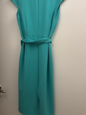 Turkos midiklänning med knytband - Snygg turkos midiklänning med v-ringning och knytband i midjan. Klänningen har korta ärmar och är tillverkad i ett mjukt, slätt tyg som ger ett elegant fall. Perfekt för dig som gillar stilrena och moderna plagg.