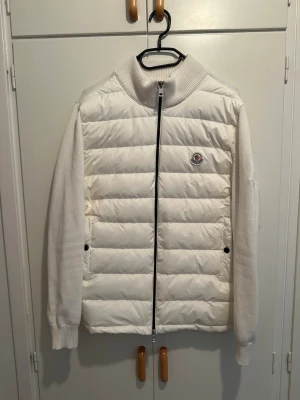 Vit Moncler cardiganjacka - Snygg vit cardiganjacka från Moncler med quiltad front och stickade ärmar samt rygg. Jackan har hög krage, dragkedja framtill och två fickor med knapp. Moncler-logga på bröstet ger en exklusiv touch. Perfekt för dig som gillar stilren och modern design. Loggan är skannigsbar.