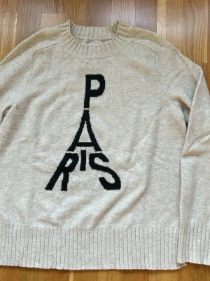 Beige stickad tröja LOFT Parismotiv - Stickad beige tröja från LOFT med svart text 'PARIS' i ett coolt grafiskt tryck på framsidan. Tröjan har rund halsringning och ribbade muddar vid ärmslut och nederkant. Perfekt för dig som gillar trendiga statement-plagg.