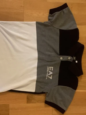 EA7 Emporio Armani pikétröja - Snygg pikétröja från EA7 Emporio Armani med färgblock i svart, grått och vitt. Tröjan har klassisk krage, två knappar och tydlig logga på bröstet. Tillverkad i mjukt bomullsmaterial som känns skönt mot huden. Perfekt för en stilren och sportig look.