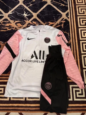 PSG träningsset Nike vit/rosa M - Snyggt PSG träningsset från Nike med långärmad vit tröja och rosa ärmar, svarta detaljer och PSG-logga på bröstet. Matchande svarta byxor med rosa och PSG-märke. Perfekt för fotbollsträning eller chill. Materialet är funktionspolyester.