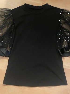 Svart topp med puffärmar och stjärnor - Cool svart topp med transparenta puffärmar i mesh dekorerade med små silverfärgade stjärnor. Ärmarna har även breda satinband vid avslutet för extra statement-look. Perfekt för dig som vill sticka ut lite extra.