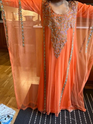  till barn 9-11år - Säljer en orange långklänning med vackra pärl- och paljettbroderier i blått och guld framtill. Klänningen har långa ärmar och transparent tyg med dekorativa detaljer längs kanterna. Perfekt för dig som vill sticka ut med en unik och elegant look.