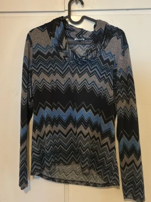 Mönstrad tröja med zigzag-mönster - Säljer en långärmad tröja med grå, blå och svarta zigzag-mönster. Tröjan har en huva och är tillverkad i ett mjukt, stretchigt material som känns skönt mot huden. Perfekt för dig som gillar unika mönster och vill sticka ut lite extra.