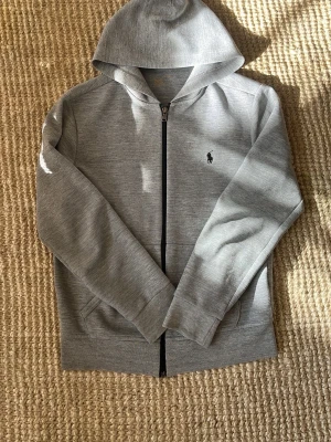Grå zip hoodie från Polo Ralph Lauren - Snygg grå hoodie med dragkedja från Polo Ralph Lauren. Klassisk design med svart broderad logga på bröstet, huva och fickor framtill. Tillverkad i mjukt bomullsmaterial som är skönt att ha på sig. Perfekt till chill eller streetwear-stil.