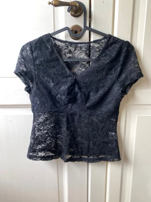 Svart spetsblus med v-ringning - Snygg svart blus i genombruten spets med v-ringad hals och korta ärmar. Figurnära passform som ger en elegant siluett. Perfekt att styla med jeans eller kjol för en trendig look.