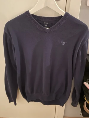 Mörkblå V-ringad tröja från Gant - Klassisk mörkblå V-ringad tröja från Gant med diskret logga broderad på bröstet. Tröjan har långa ärmar, ribbade muddar och är tillverkad i mjukt material som passar perfekt till lager-på-lager-stil.