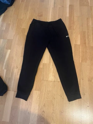 Svarta mjukisbyxor från BOSS - Snygga svarta mjukisbyxor från BOSS med broderad logga på benet. Byxorna har dragsko i midjan och ribbade muddar vid bensluten. Perfekta för chill dagar och har en klassisk, enkel design som funkar till det mesta.