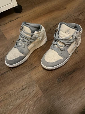 Nike Air Jordan 1 Mid grå/vit - Snygga Nike Air Jordan 1 Mid sneakers i grått och vitt. Skorna har klassisk hög siluett, grå mockadetaljer och vita paneler i textil. Ikoniska Nike-loggan på sidan och Air Jordan-vingen vid ankeln. Perfekta för dig som gillar streetwear och sportig stil. Storlek 38