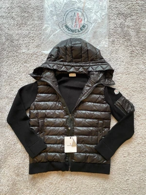 Svart pufferjacka från Moncler - Den är L men kan passa beroende på vikt och längd, hör av er via intresse. Snygg svart pufferjacka från Moncler med stickade ärmar och huva. Jackan har quiltad front i glansigt material, dragkedja och två fickor fram. Moncler-logga på ärmen och ribbade muddar för extra komfort. Perfekt för kalla vinterdagar. 