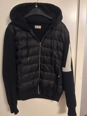 Monclear cardigan  - Snygg svart dunväst från Moncler med stickade ärmar och stor huva. Västen har dragkedja framtill, två fickor med knappar och ett vitt zigzag-mönster på ena ärmen samt Moncler-logga. Perfekt för lager på lager och streetwear-stil.