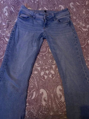 Ljusblå bootcut jeans med brodyr - Säljer ett par ljusblå bootcut jeans med snygg brodyr på bakfickorna och knappar på ficklocken. Klassisk femficksmodell i mjukt denimtyg, perfekta till en avslappnad stil. Passar dig som gillar detaljer och vill ha något extra i garderoben. Lite slitna längst ner men inget som påverkar utseendet på jeansen.