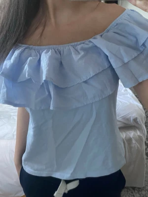 Ljusblå offshoulder topp med volang - Supersöt ljusblå offshoulder topp med dubbla volanger runt hela axelpartiet. Toppen är ärmlös och har en luftig passform, perfekt för varma dagar. Tillverkad i mjuk bomull som känns skön mot huden.