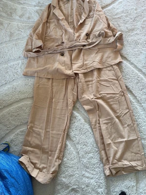 Beige kostym från SHEIN Curve 1XL - Säljer en beige kostym från SHEIN Curve i storlek 1XL. Kostymen består av en kavaj med knappar och tillhörande bälte samt matchande byxor med uppvikta benslut. Materialet känns lätt och passar perfekt för en stilren look.