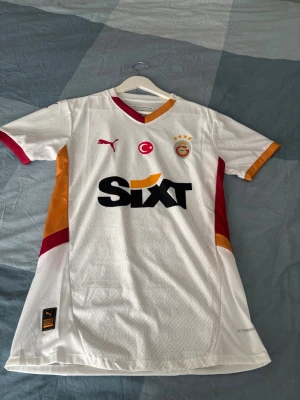 Galatasaray vit matchtröja Puma - Snygg vit Galatasaray matchtröja från Puma med röda och orange detaljer. Tröjan har klubbmärke, Puma-logga och sponsortryck på bröstet samt mönstrad struktur i tyget. Perfekt för fotbollsträning eller att bära till vardags. Storlek mer som Small 