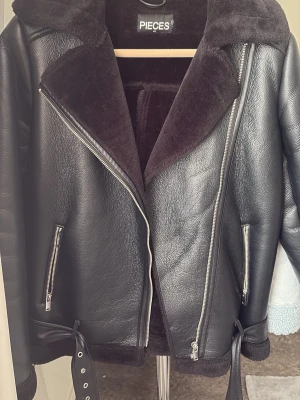 Svart biker-kappa från PIECES - Snygg svart biker-inspirerad kappa från PIECES med breda slag i fuskpäls, dragkedjor och bälte i midjan. Kappan har en cool, oversized look och är fodrad med mjuk fuskpäls för extra värme. Perfekt för dig som vill ha en edgy stil.