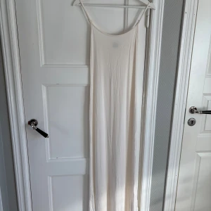 Lång vit sommarklänning med tunna band - Elegant långklänning i vitt med tunna axelband och enkel, rak siluett. Perfekt för varma dagar och har en minimalistisk design som ger en stilren look. Tillverkad i ett mjukt, lätt material som känns svalt mot huden.