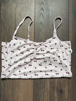 Ljusrosa linnetopp med körsbärsmönster - Söt ljusrosa linnetopp med tunna axelband och rynkdetalj framtill. Toppen har ett gulligt körsbärsmönster i rött och grönt och är gjord i mjuk bomull. Perfekt för varma dagar och enkel att matcha med jeans eller kjol.