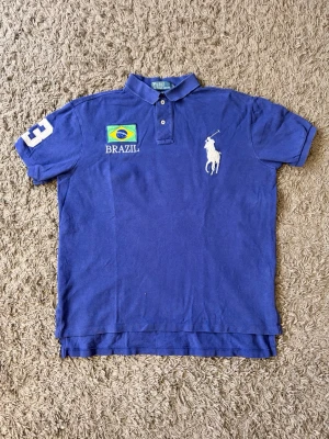 Chief Keef Ralph Lauren Brazil piké big pony - Säljer en blå piké från Ralph Lauren med stor vit logga på bröstet och Brasilien-flagga med texten BRAZIL. Tröjan har korta ärmar och nummer 3 på ärmen. Klassisk krage och knappar framtill. Perfekt för dig som gillar sportig stil.