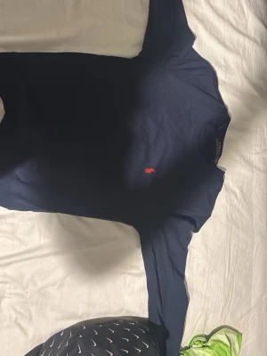 Mörkblå tröja från Ralph Lauren - Klassisk mörkblå tröja från Ralph Lauren med rund halsringning och den ikoniska röda logotypen broderad på bröstet. Tröjan har långa ärmar och är tillverkad i ett mjukt, tunt material som passar perfekt till lager på lager.