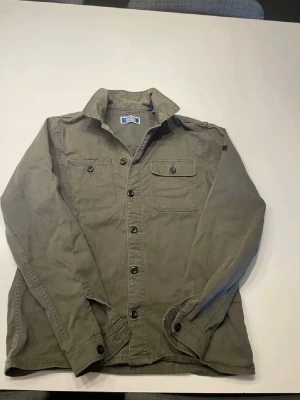 Olivgrön overshirt från Jack and Jones - Stilren olivgrön overshirt från Jack and Jones med två bröstfickor med lock och knappar, klassisk krage och svarta knappar längs hela framsidan. Tillverkad i ett mjukt bomullstyg som ger en avslappnad look. Perfekt att bära öppen över en t-shirt.