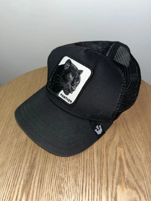 Svart Goorin Bros Panther - Svart truckerkeps från Goorin Bros med mesh baktill och böjd skärm. Framtill sitter ett broderat patch med en svart panter och texten 'Panther'. Kepsen har justerbar snapback och är tillverkad i bomull och polyester. | Har du frågor eller funderingar? Skicka PM‼️